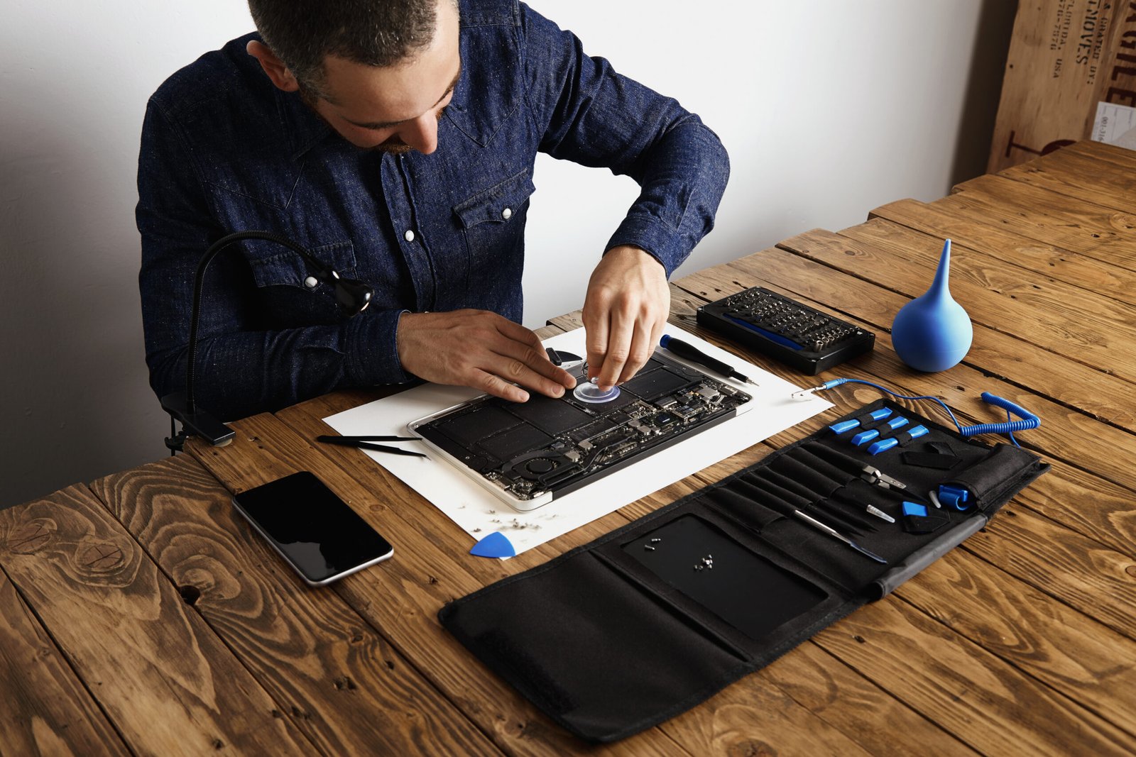 Using small suction cup to remove battery cells from broken laptop to repair and clean it in his laboratory with special tools on table around.